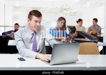 Mid adult businessman sitting at desk, using laptop, colleagues working in background Stock Photo