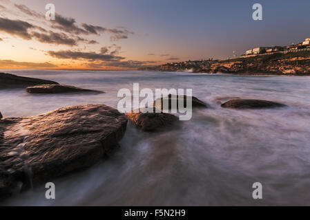 Seascape sunrise long exposure with flowing water around large rocks and light pink and blue sky Stock Photo