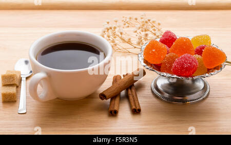 A fresh cup of hot tea, colorful marmalade, brown sugar cane cubes and cinnamon sticks..Close-up on wooden table Stock Photo