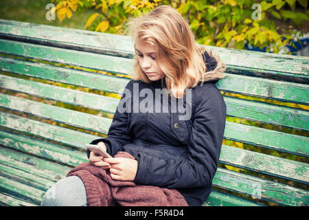 Beautiful Caucasian blond teenage girl in black jacket sitting on old green park bench with smart phone in hands Stock Photo