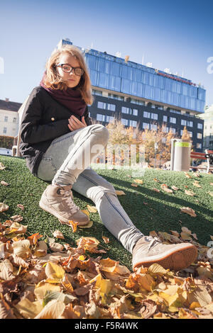 Beautiful Caucasian blond teenage girl in glasses sitting in autumnal city park with falling leaves Stock Photo