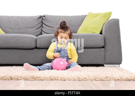 Cute little baby girl putting a coin into a pink piggybank seated in front of a gray sofa isolated on white background Stock Photo