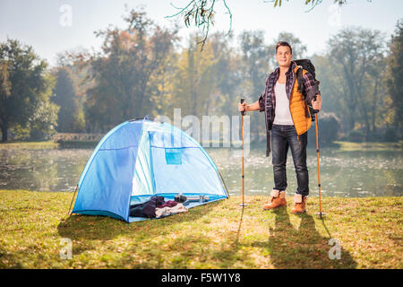 Young male hiker posing by a pond next to a blue tent in a field on a sunny autumn day Stock Photo