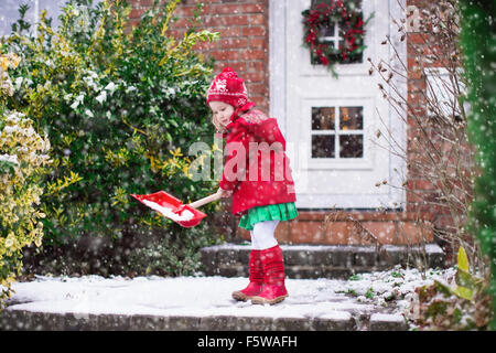 Little girl shoveling snow on home drive way. Beautiful house decorated for Christmas. Child with shovel playing outdoors Stock Photo