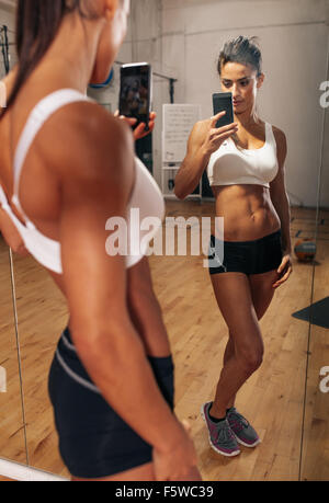 Fitness model taking a self portrait from the reflection in the mirror using her mobile phone. Muscular young woman making selfi Stock Photo