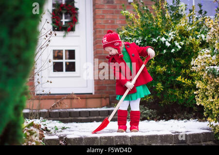 Little girl shoveling snow on home drive way. Beautiful house decorated for Christmas. Child with shovel playing outdoors Stock Photo