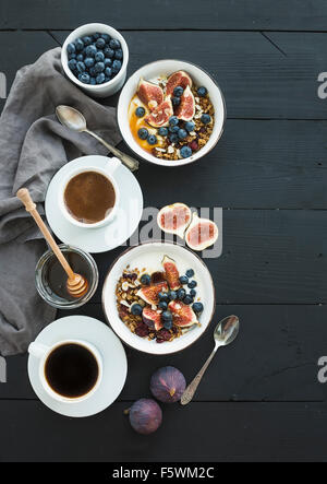 Healthy breakfast set. Bowls of oat granola with yogurt, fresh blueberries and figs, coffee, honey, over black wooden backdrop. Stock Photo