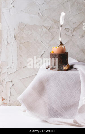 Breakfast with started eating soft-boiled egg with pouring yolk in wooden eggcup with silver spoon on white cloth over white tab Stock Photo