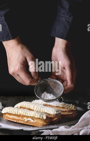 Eclairs with white butter cream in vintage metal plate with Sifting sugar powder from woman's hands. Dark rustic style. Day ligh Stock Photo