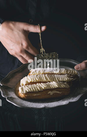 Woman's hands take by cake tongs from vintage metal plate Eclairs with white butter cream. Dark rustic style. Day light. In retr Stock Photo