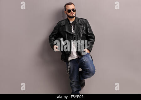 Young male biker in black leather jacket smoking a cigarette and leaning against a gray wall Stock Photo