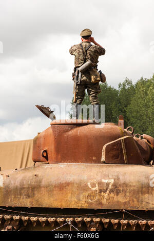 World war two re-enactment. German officer (seen from behind), standing on burned out Sherman tank, observing through binoculars. Stock Photo
