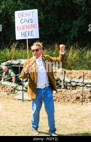 Vietnam War re-enactment. Lone Peace-movement protester standing and shouting with placard "Draft card burning here," in front of trench and soldier. Stock Photo