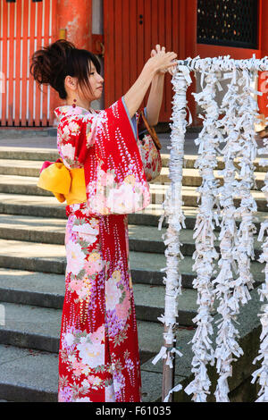 Side view of young Japanese woman in red kinomo reaching up to tie omikuji, a paper fortune slip to framework already filled with thousands of slips. Stock Photo