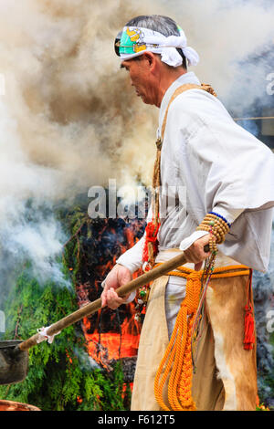 Japan, Nishinomiya, Mondo Yakuji temple. Priest in yamabushi costume ladling cups of scared water over smoking ritual bonfire during monthly ceremony. Stock Photo