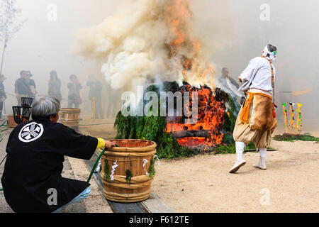 Japan, Nishinomiya, Mondo Yakuji temple. Priest in yamabushi costume ladling cups of scared water over smoking ritual bonfire during monthly ceremony. Stock Photo