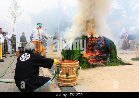 Japan, Nishinomiya, Mondo Yakuji temple. Priest in yamabushi costume ladling cups of scared water over smoking ritual bonfire during monthly ceremony. Stock Photo