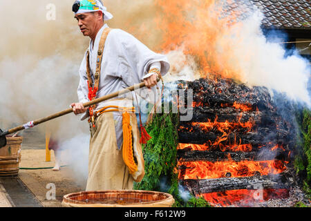 Japan, Nishinomiya, Mondo Yakuji temple. Priest in yamabushi costume ladling cups of scared water over smoking ritual bonfire during monthly ceremony. Stock Photo