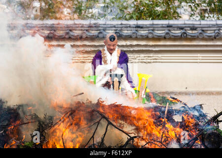 Japan, Nishinomiya, Mondo Yakuji, Shotaizan Tokoji temple. High Priest in yamabushi costume sitting behind ritual bonfire. Heat haze distortion. Stock Photo