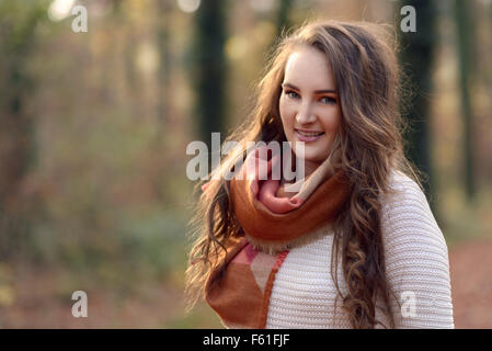 Pretty fashionable young woman standing outdoors in an autumn forest wearing a chic scarf over her jersey smiling at the camera,