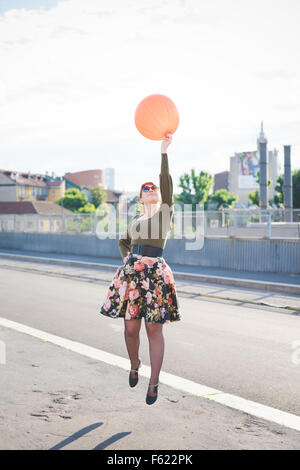 young handsome caucasian redhead woman jumping, playing with orange balloon, hand on her hips, looking up - youth, childhood concept - wearing floral skirt and green shirt Stock Photo