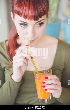 Half length of young handsome caucasian redhead woman sitting in a bar, drinking a juice with a straw, looking downward - relaxing, happy hour concept - wearing green shirt Stock Photo