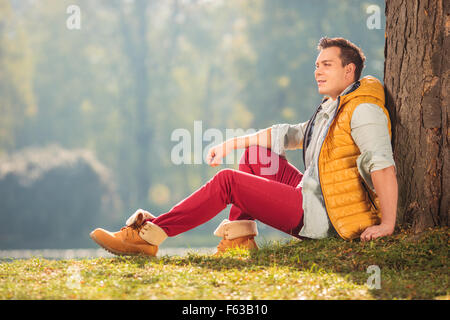 Joyful young man sitting on the grass in a park and leaning against a tree on a beautiful autumn day Stock Photo