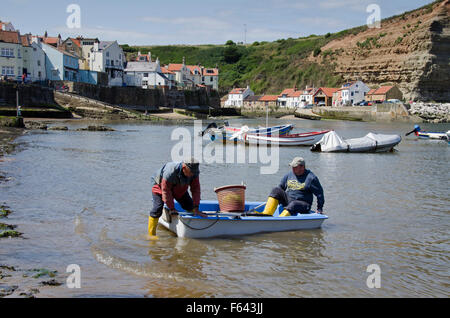 2 fishermen in a small boat, land their catch in a bucket on the sea shore - sunny summer day, harbour beach, Staithes' village, North Yorkshire, UK. Stock Photo