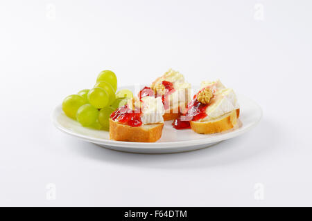 slices of fresh bread with brie cheese, walnuts, jam and grapes on white plate Stock Photo