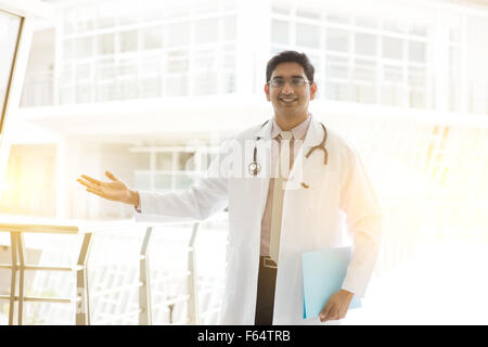 Portrait of a smiling Asian Indian male medical doctor standing inside hospital, holding file folder and showing welcome hand si Stock Photo