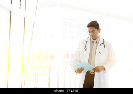 Portrait of handsome Asian Indian medical doctor walking and reading on medical report, outside hospital building block, beautif Stock Photo