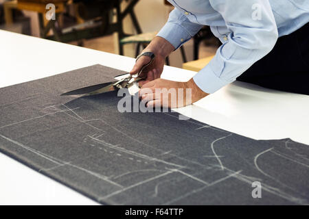 Tailor working in his shop cutting a roll of dark fabric on which he has marked out the pattern of the garment he is making with Stock Photo