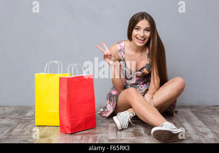 Pretty excited cheerful young woman with long hair sitting on the floor near colorful gifts and showing peace sign Stock Photo