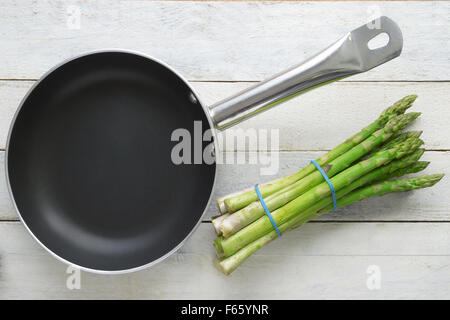 Bunch of fresh asparagus next to a frying pan on a white wooden table. Stock Photo
