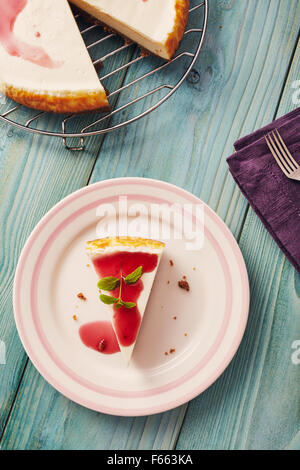 new york cheesecake with cherry sauce on a plate on a blue wooden table, full cake in background Stock Photo