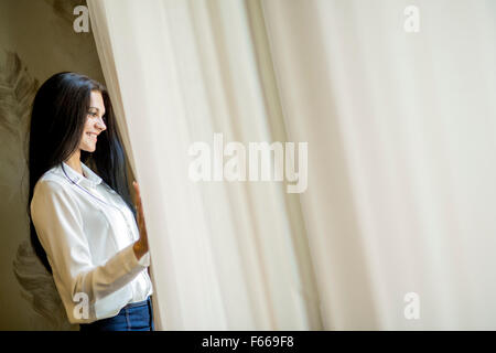 Happy beautiful young woman staring out the window and holding curtains Stock Photo