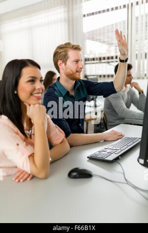 Beautiful young woman and a man raising hands in classroom in  while sitting in fron of a desktop Stock Photo