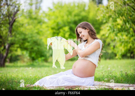 Beautiful pregnant woman holding baby bodysuit and smiling outside in the park Stock Photo