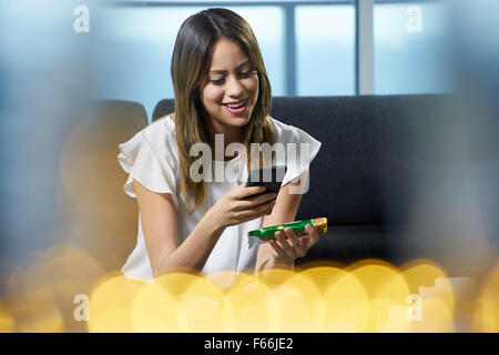 Young hispanic woman at home, holding a cereal bar and reading nutritional facts on package. She uses an app on her phone to sca Stock Photo