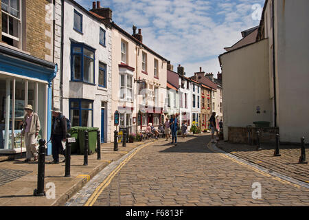 People sit by a pub or take a summer stroll down the sunny cobbled High Street - quaint fishing village of Staithes, North Yorkshire, England, UK.