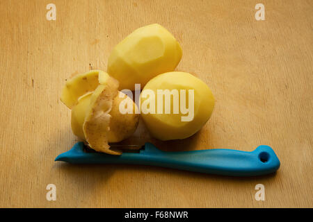 Three potato lying on an old wooden table, two of them are peeled and the third is chosen.partly. Next is blue potato peeler, wi Stock Photo