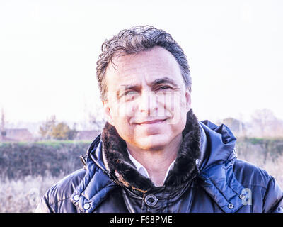 Elegant middle-aged man with green eyes and salt pepper hair dressed in dark blue padded jacket is smiling reassuringly in the countryside Stock Photo