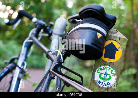 As weather warms, more people enjoy walking and cycling in London's parks and walkways. A Specialized road style bicycle with a pannier rack and saddle bag is seen propped up against a Capital Ring direction marker on a green trail across the city. Stock Photo