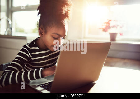 Young African girl amusing herself at home sitting at the dining table browsing the internet on a laptop computer Stock Photo