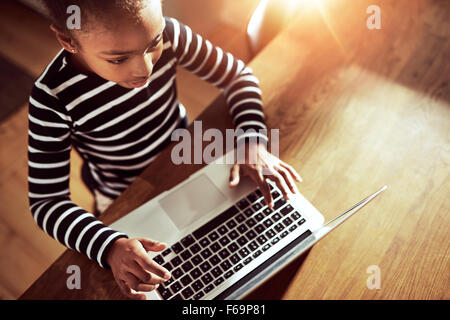 Young ethnic black girl sitting at the dining table at home typing on a laptop computer, high angle view looking down on the key Stock Photo