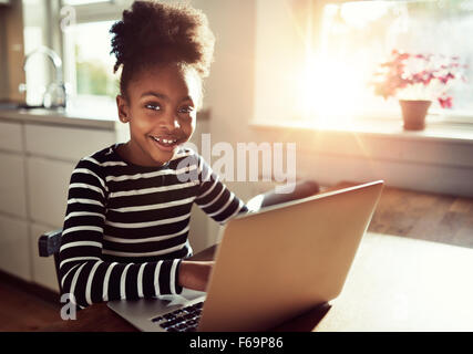 Smiling friendly young black african girl with a cute top knot afro hairstyle sitting at a laptop computer at the dining table g Stock Photo