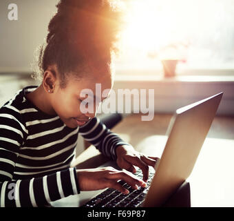 young black girl with a fun afro topknot concentrating as she types on a laptop computer, close up side view with sun flare back Stock Photo