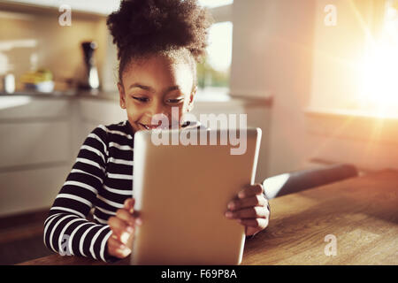 Happy little ethnic black girl sitting at home in the kitchen reading on a tablet computer grinning with pleasure, bright sun gl Stock Photo