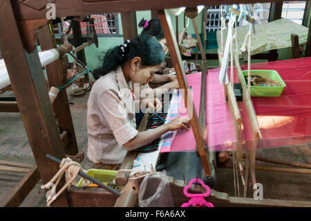 Two women weaving on a horizontal loom using shuttles with thread spools. Partially woven silk fabric visible. Mandalay, Myanmar Stock Photo