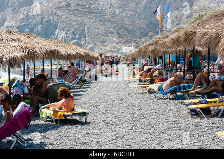 Santorini, Thira. Telephoto shot along Kamari beach with rows of sun recliners, parasols, with sunbathers and tourists, rocky cliff face as background. Stock Photo